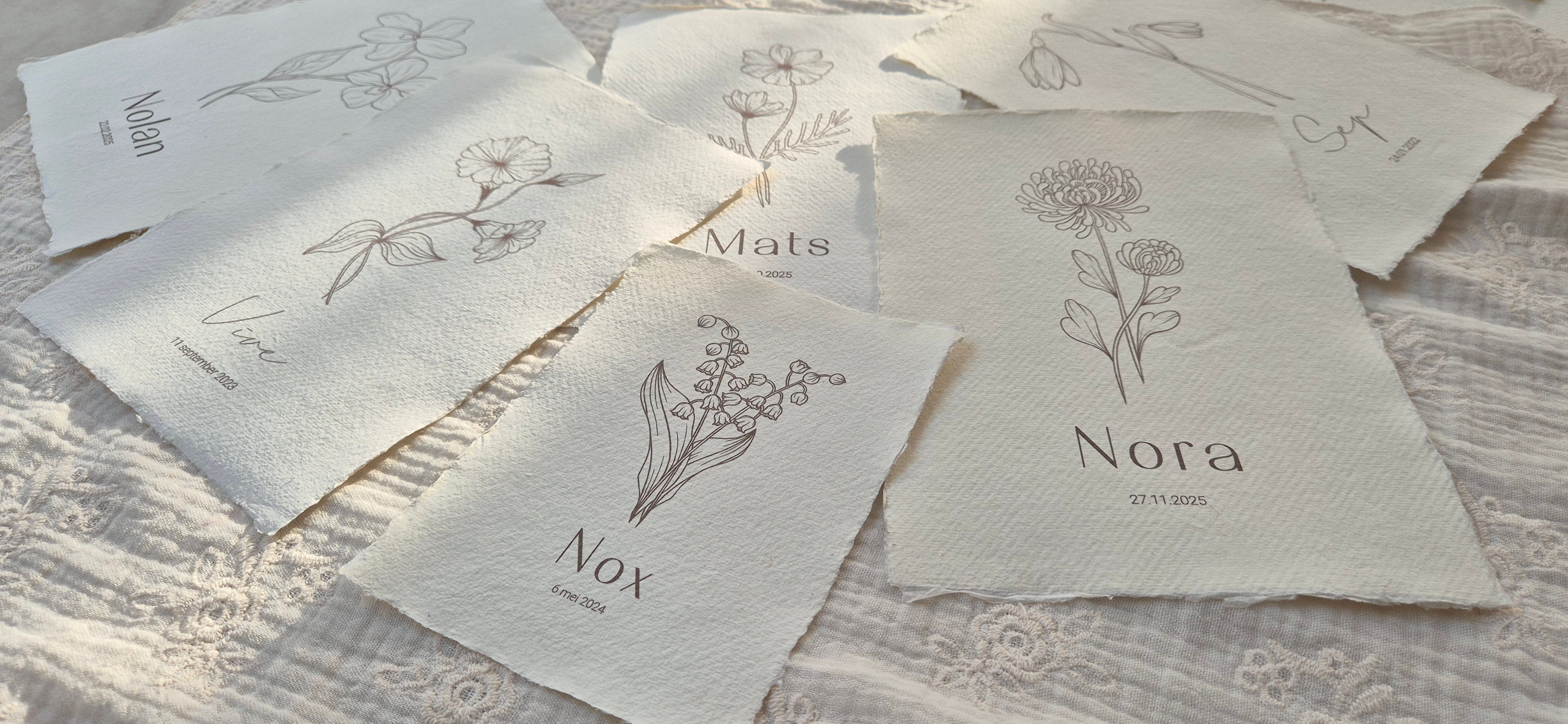 Handgemaakte geboortebloem posters op geschept papier met ruwe randen, verspreid over een zachte, lichte ondergrond. Elke poster toont een fijne botanische lijnillustratie met daaronder een naam en geboortedatum. De natuurlijke tinten en het ambachtelijke papier zorgen voor een rustige, tijdloze uitstraling.