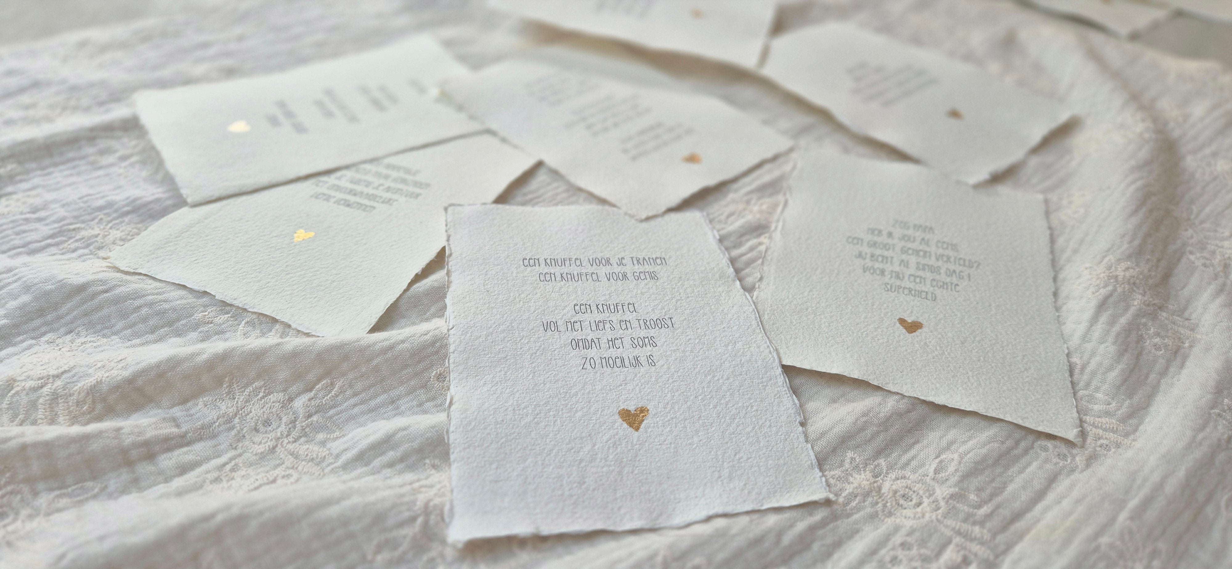 Handgemaakte gedichtposters op geschept papier met ruwe randen, los verspreid over een zachte, lichte stoffen ondergrond. Elke poster bevat een liefdevolle Nederlandse tekst in fijne typografie, afgewerkt met een klein goudkleurig hartje. De natuurlijke materialen en rustige tinten geven een warme, serene en ambachtelijke uitstraling.