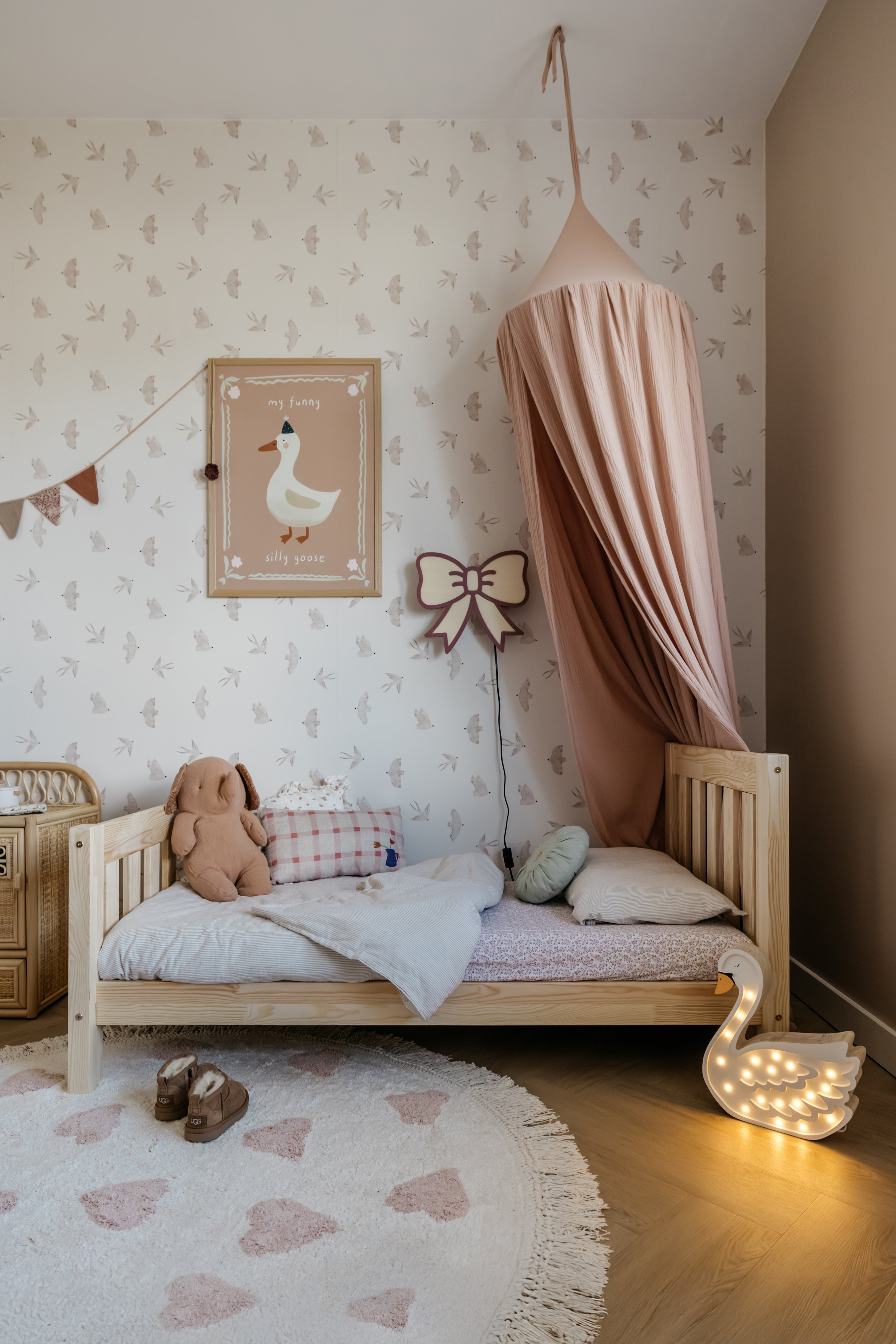 Sfeervolle babykamer met zacht patroonbehang, houten bedje met roze hemel, knuffels en warme accessoires in rustige natuurlijke tinten.