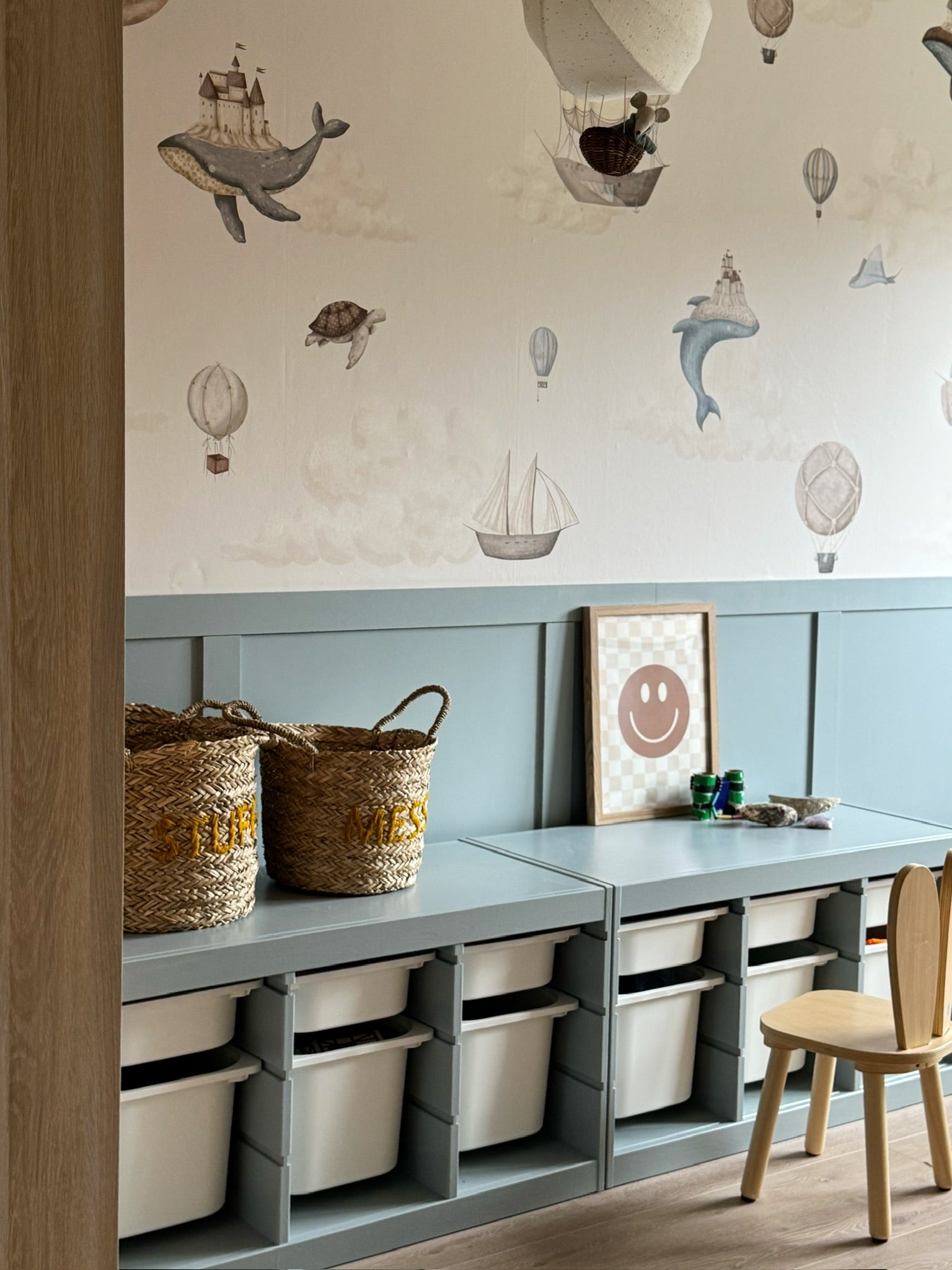 Kinderkamer met Memories of Dreams behang met dromerige illustraties van luchtballonnen, dieren en schepen boven zachtblauwe lambrisering en opbergmeubel.